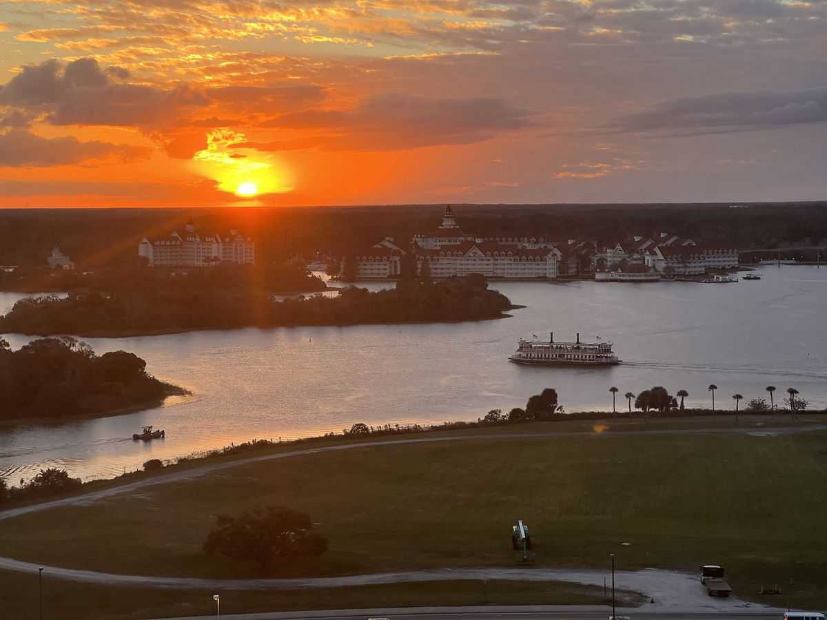 Golden sunset shimmers over a serene Disney World lake, where boats glide past iconic resort buildings. Lush trees and manicured grass frame the view, capturing that unmistakable Disney magic—every evening sparkling with the promise of unforgettable memories, like sipping their legendary $137 cocktail.