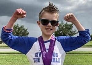 Young World Changer beaming with pride, rocking sunglasses and flexing superhero-style, shows off a hard-earned purple medal in the vibrant Give Kids the World Village—capturing the magic and celebration of an unforgettable theme park experience.