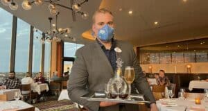A staff member in a sharp suit and blue mask brings drinks to guests on a tray inside the elegant, window-lined restaurant at Universal Studios.