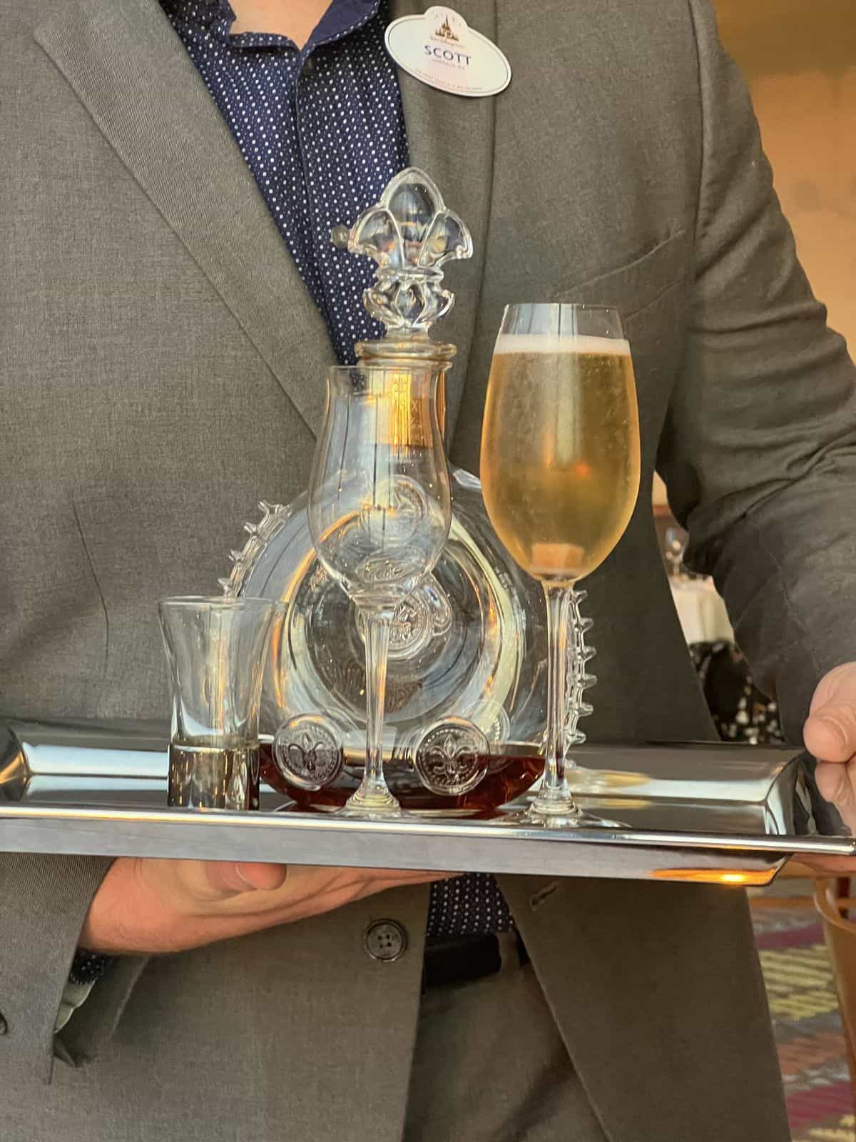 A sharply dressed server at Walt Disney World gracefully presents a tray adorned with a decanter, a cocktail shot glass, and a sparkling champagne flute, embodying the sophisticated atmosphere of Disney’s most luxurious lounges.