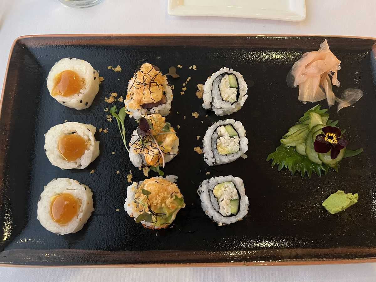 A vibrant assortment of sushi rolls on a black plate, joined by pickled ginger, wasabi, and a whimsical cucumber flower garnish—an ideal culinary adventure to enjoy alongside one of Walt Disney World's legendary signature cocktails.