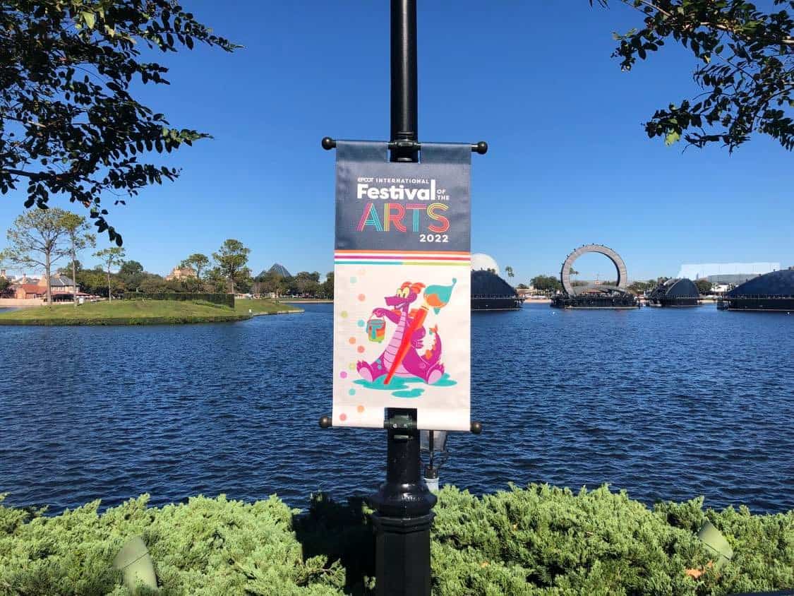 EPCOT International Festival of the Arts 2022 banner on a lamppost by World Showcase Lagoon, surrounded by lush trees and bright blue skies, inviting guests to celebrate the color and creativity of the Performing Arts at EPCOT.