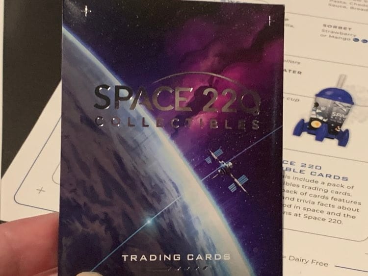 A fan shows off a Space 220 Collectibles trading card, spotlighting the iconic space station against the restaurant’s signature purple-blue cosmic backdrop, capturing the immersive vibe of EPCOT’s Space 220 dining experience.