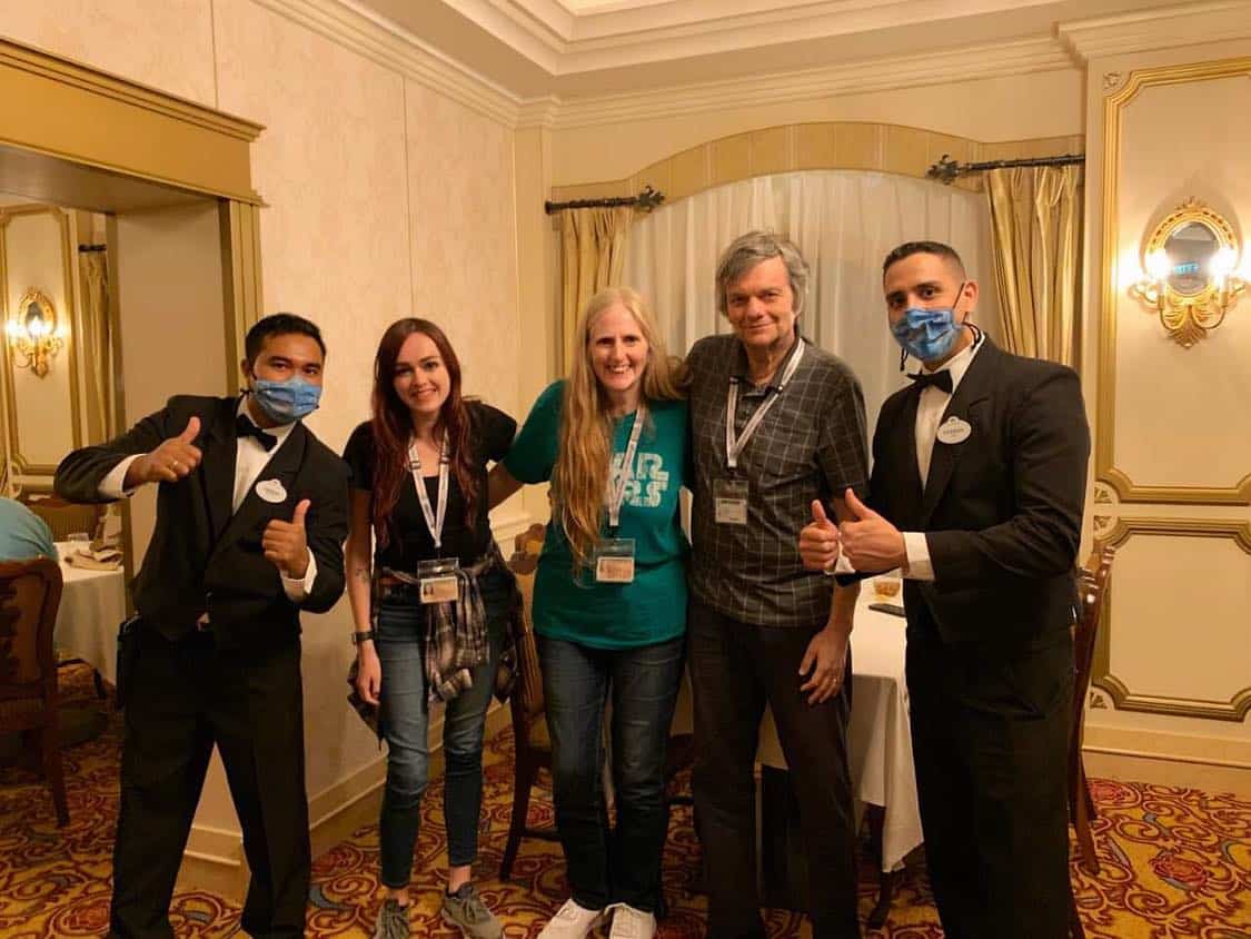 A group of five guests beams for a photo in an elegantly decorated Disney Cruise Line dining room, glowing with warm light. Two are dressed in formal black suits with blue face masks, evoking the signature look of Disney Cruise Line Crew Members who ensured onboard safety during the Covid era.