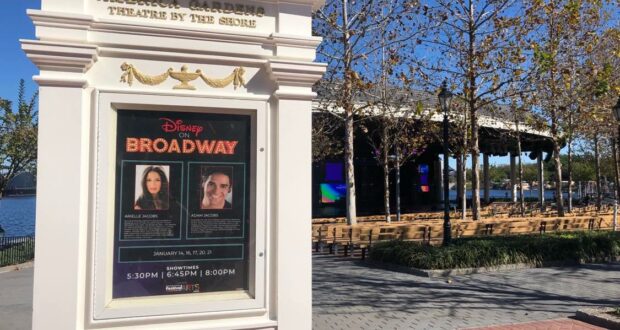 Disney Broadway concert poster displayed on a white sign near the outdoor stage at EPCOT, surrounded by empty audience seats and lush trees, inviting guests to experience the vibrant energy of the EPCOT International Festival of the Arts and its celebration of live performances.