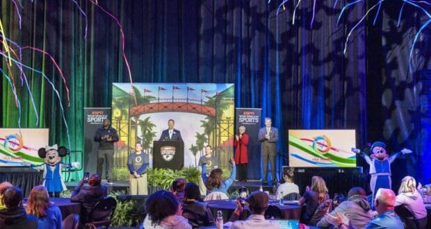Guests gather in a vibrant Disney World theater to watch a special event where Disney Live Entertainment teams up with Special Olympics USA. The stage features beloved characters, energetic speakers, colorful streamers, and festive decorations filling the venue.