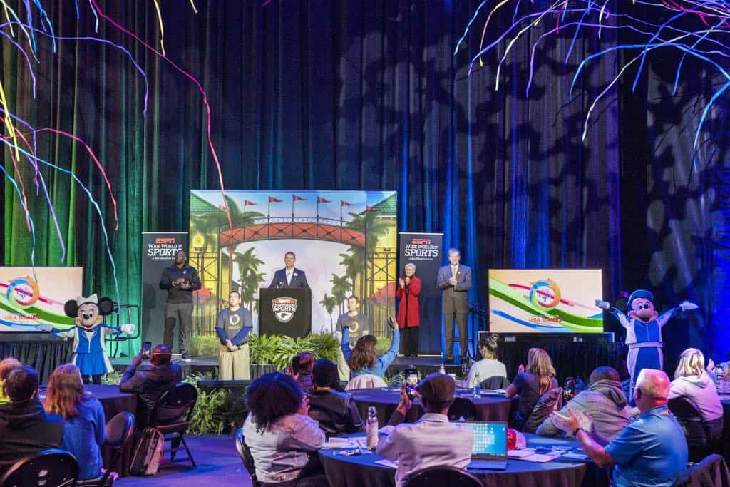 Guests gather in a vibrant Disney World theater to watch a special event where Disney Live Entertainment teams up with Special Olympics USA. The stage features beloved characters, energetic speakers, colorful streamers, and festive decorations filling the venue.