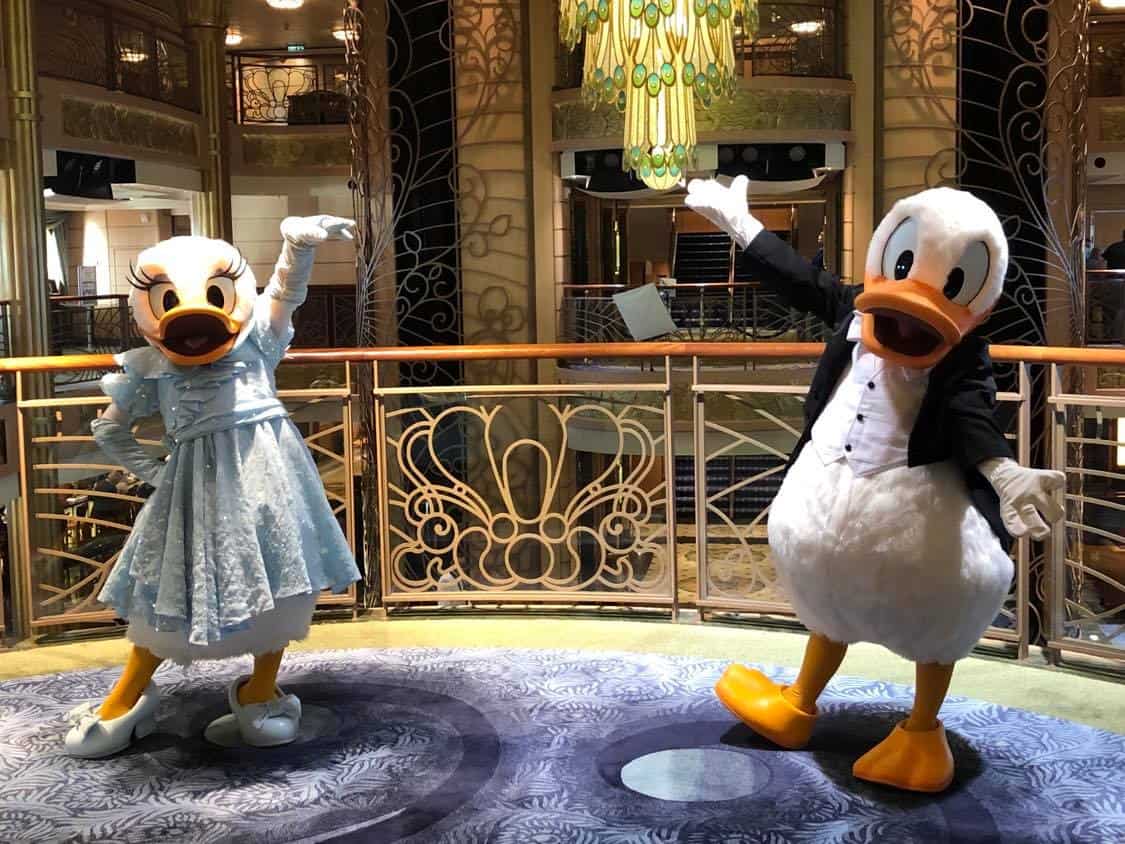 Donald Duck and Daisy Duck, dressed in their signature outfits, greet guests from a lavish ship balcony adorned with sparkling chandeliers aboard Disney Cruise Line, creating an enchanting moment for fans during the unique experiences of sailing in the Covid era.