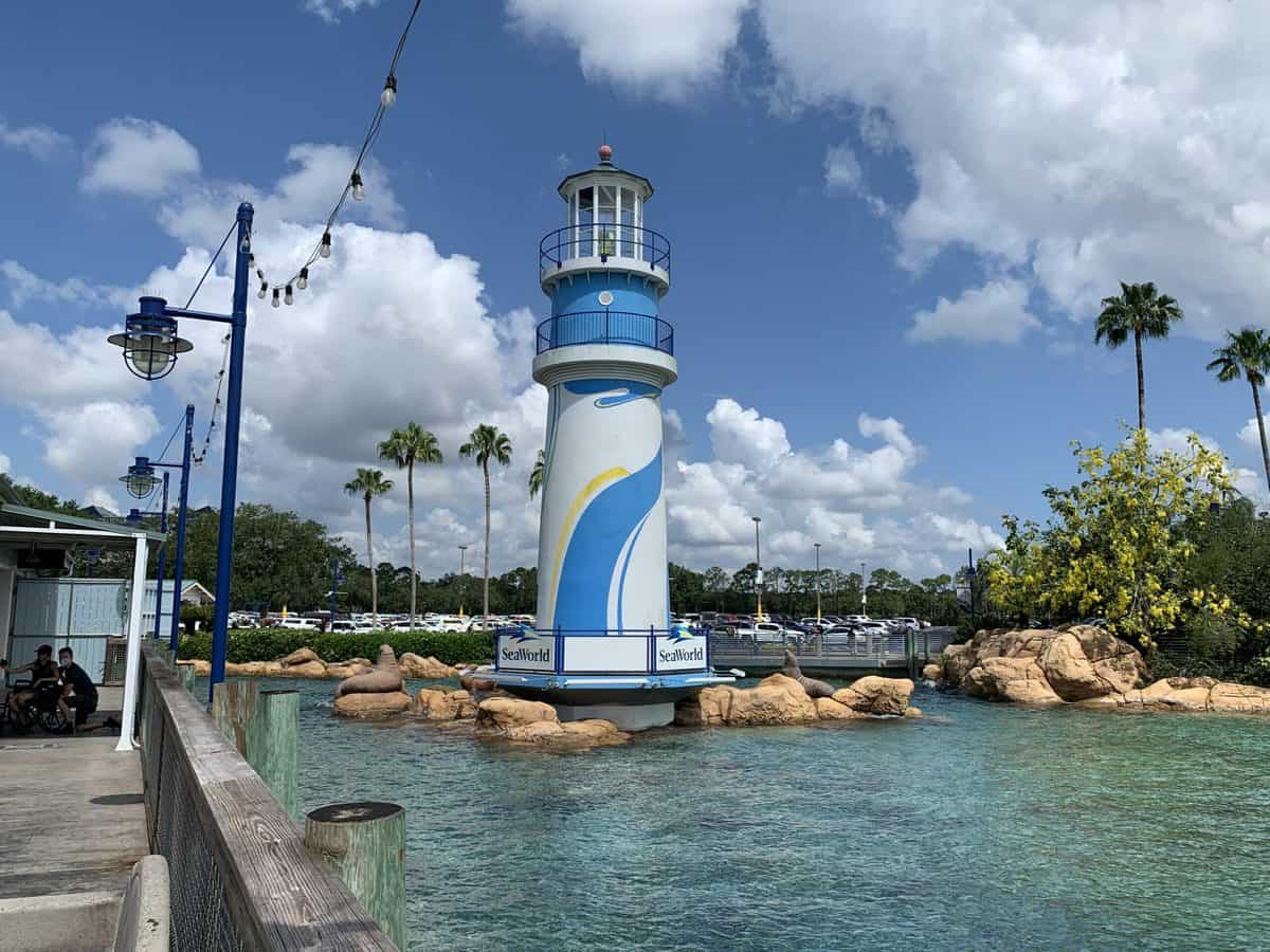 The scenic white lighthouse with blue accents at this Florida attraction rises beside sparkling water, flanked by rocks and swaying palm trees beneath a sunny sky—a must-see photo spot where preschoolers enjoyed free admission in 2022.