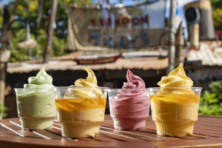 Four cups of vibrant soft serve ice cream on a wooden table at Disney’s Typhoon Lagoon, surrounded by lush greenery, perfectly embodying the playful atmosphere and excitement of this iconic water park.