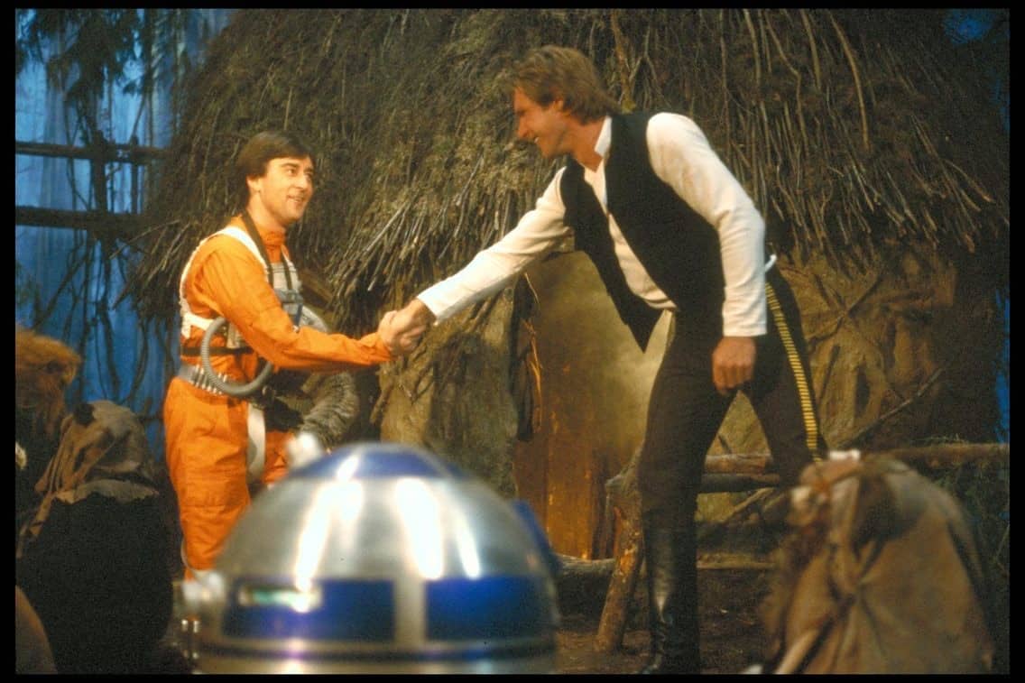 Guests experience a Star Wars-themed attraction with two characters shaking hands outside a rustic hut, while R2-D2 and Ewoks interact nearby in a forested area inspired by scenes from The Book of Boba Fett.