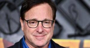 A cheerful guest with brown hair and glasses poses for a photo, wearing a blue shirt and dark jacket, as entertainment news about The Family of Bob Saget's lawsuit over death records flashes across the TV screens in the park’s lounge area.