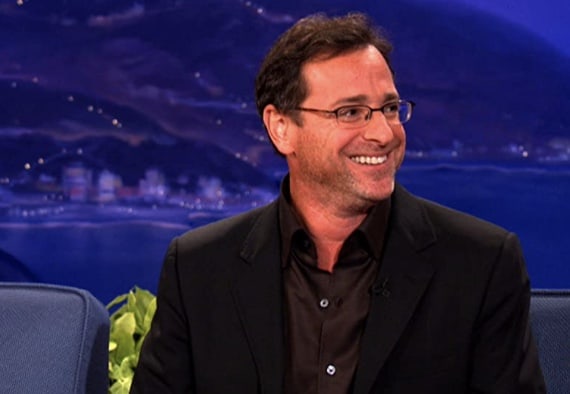 TV host in glasses and a sharp black suit beams on stage, framed by the vibrant Orlando skyline, channeling the memorable energy of Bob Saget’s iconic Tonight Show moments—perfectly capturing the magic and excitement of Orlando’s entertainment scene.