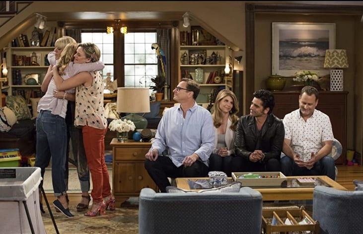 Six friends gather in a welcoming Orlando living room, sharing laughter and heartfelt stories about the legendary Bob Saget; two women embrace while others smile warmly, cherishing their unforgettable memories from the world of comedy and entertainment.