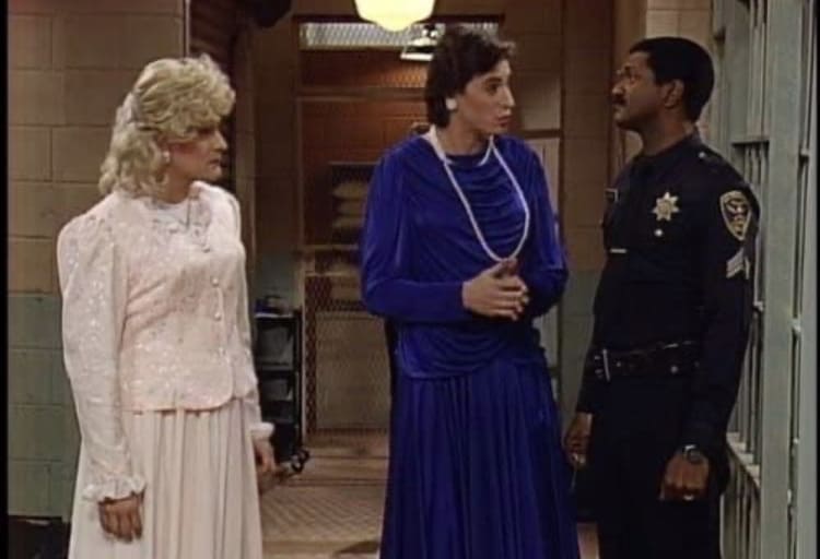 Two performers dressed in elaborate gowns chat with a uniformed police officer in a hallway lined with beige walls and metal bars, evoking the whimsical energy of an Orlando stage show that once featured Bob Saget in a fan-favorite role.
