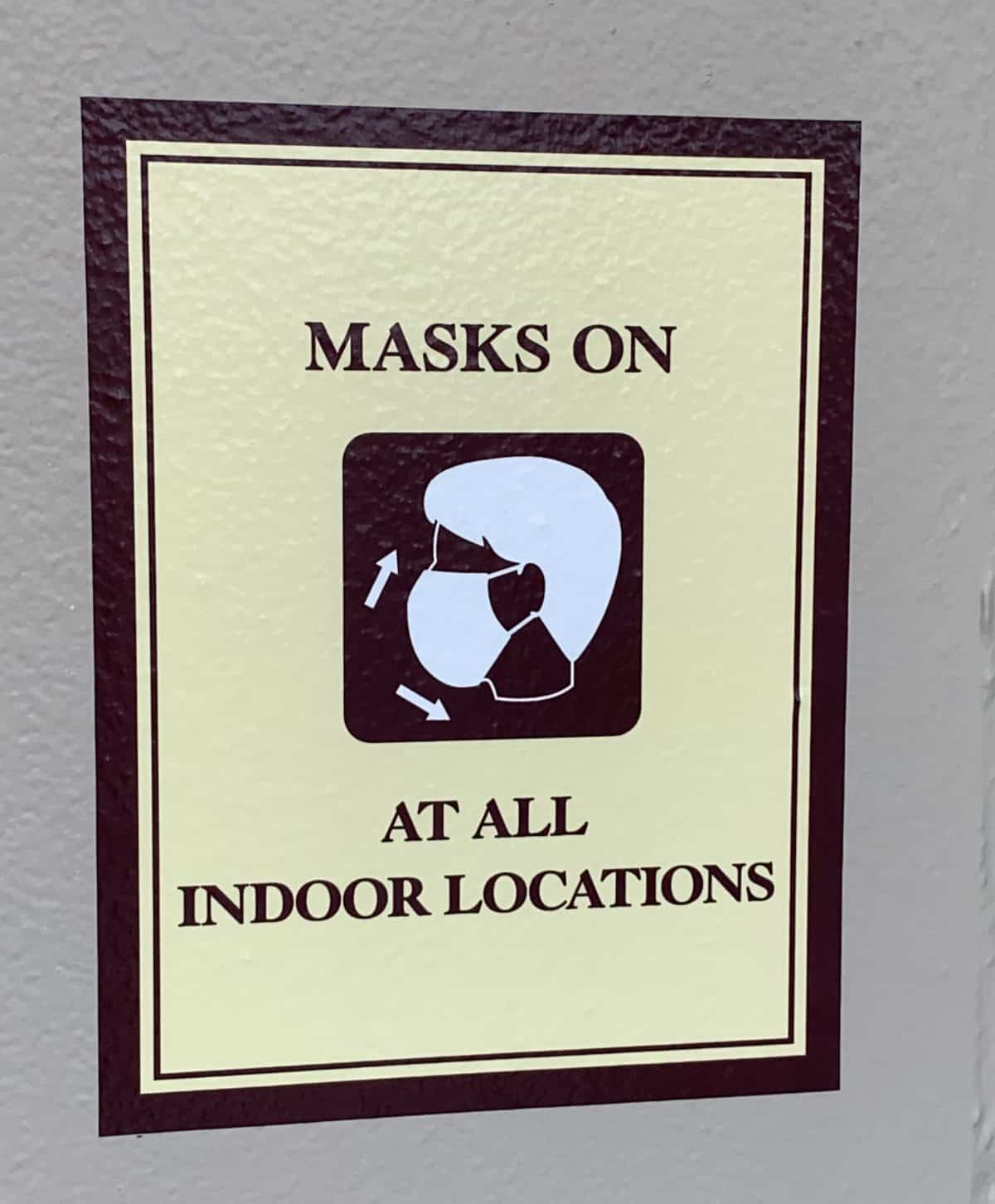 Sign at Typhoon Lagoon Disney Water Park displays: "Masks on at all indoor locations" alongside an icon of a guest wearing a face mask, reminding visitors that complimentary face coverings are offered for your safety and convenience.