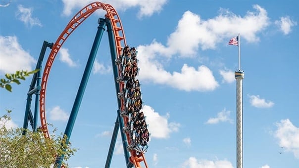 Thrill-seekers plunge down the daring drop of Central Florida’s newest roller coaster, racing past a towering structure crowned with an American flag, all set against a clear blue sky for the ultimate theme park adrenaline rush.