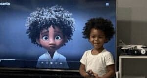 A cheerful toddler poses next to a TV screen featuring Mirabel from Disney’s Encanto, both showcasing matching curly hairstyles.
