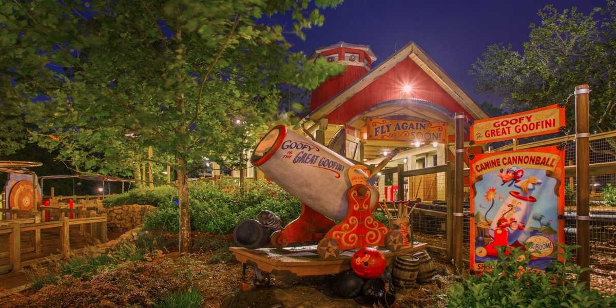 Brightly lit amusement park area at night featuring a red and white cartoonish cannon labeled "Goofy's Great Goofini" surrounded by vibrant signs, trees, and a colorful building in the background. Not far from Space Mountain in the Magic Kingdom, signs advertise "The Great Goofini" and "Cannonball.