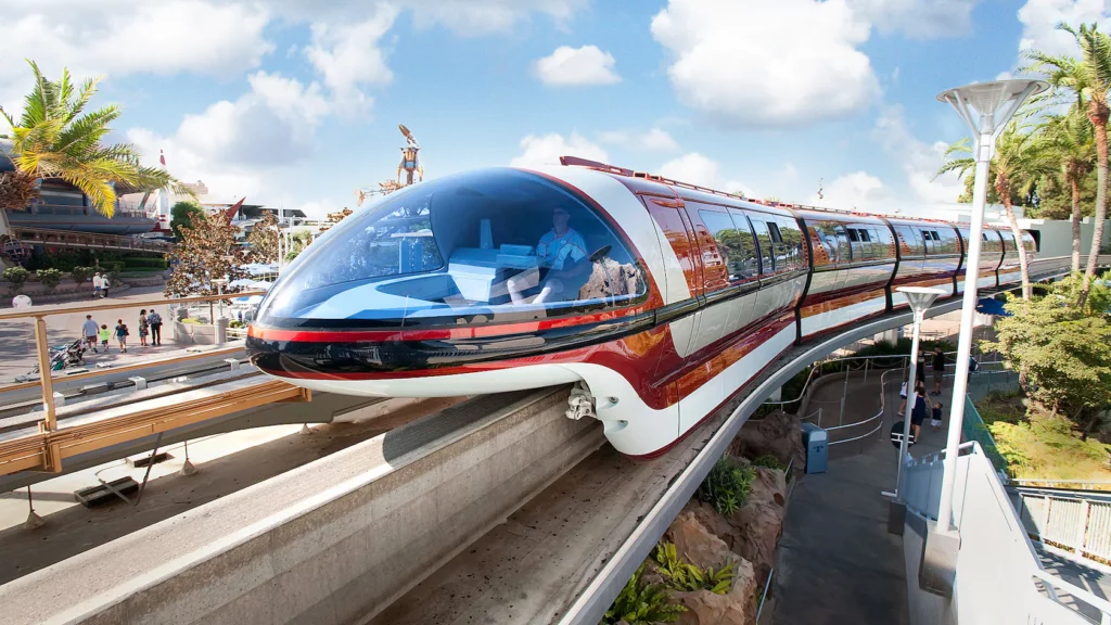 A sleek, futuristic monorail with a shiny red and white exterior travels on an elevated track under a clear blue sky. It passes through a scenic area with palm trees and modern architecture visible in the background.