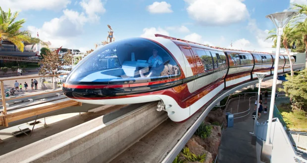A sleek, futuristic monorail with a shiny red and white exterior travels on an elevated track under a clear blue sky. It passes through a scenic area with palm trees and modern architecture visible in the background.