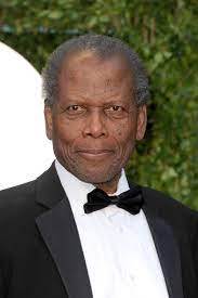 A dignified gentleman in a sharp tuxedo and classic bow tie poses outdoors with a warm, inviting smile, evoking the elegance of legendary Hollywood icon Sidney Poitier, whose legacy lives on after his passing at 94.