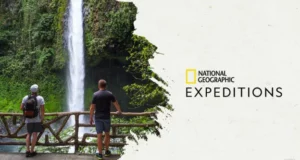 Adventurers take in the awe-inspiring view of a towering waterfall from behind a wooden railing, experiencing a thrilling National Geographic Expeditions journey in 2023. The iconic “National Geographic Expeditions” logo is visible.