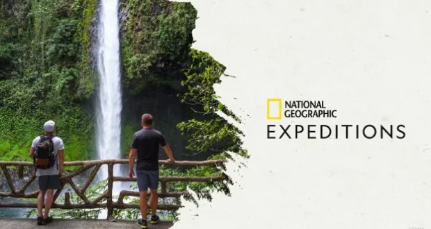 Adventurers take in the awe-inspiring view of a towering waterfall from behind a wooden railing, experiencing a thrilling National Geographic Expeditions journey in 2023. The iconic “National Geographic Expeditions” logo is visible.