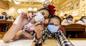 A woman and child wearing masks smile and hold pens in a lively classroom filled with other students, capturing the joy and community spirit that Disney brings to fans both inside the parks and through its outreach programs.