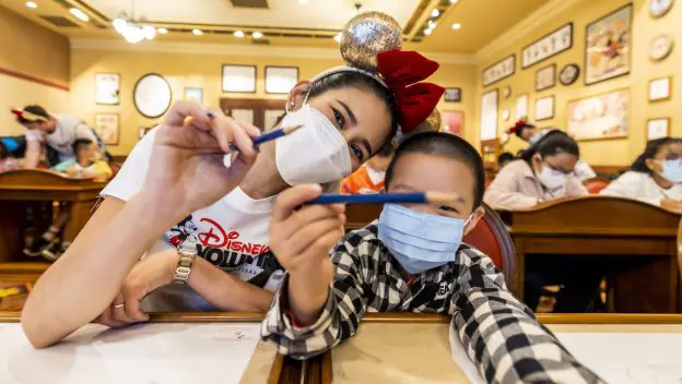 A woman and child wearing masks smile and hold pens in a lively classroom filled with other students, capturing the joy and community spirit that Disney brings to fans both inside the parks and through its outreach programs.