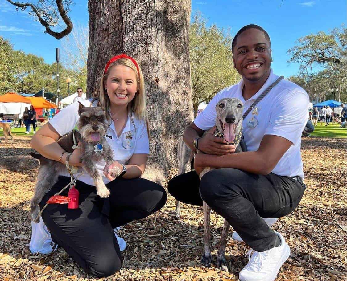At a lively Disney event, two guests kneel outdoors smiling and holding their dogs, surrounded by trees and colorful festival tents, as the celebration raises thousands to support animals in need.