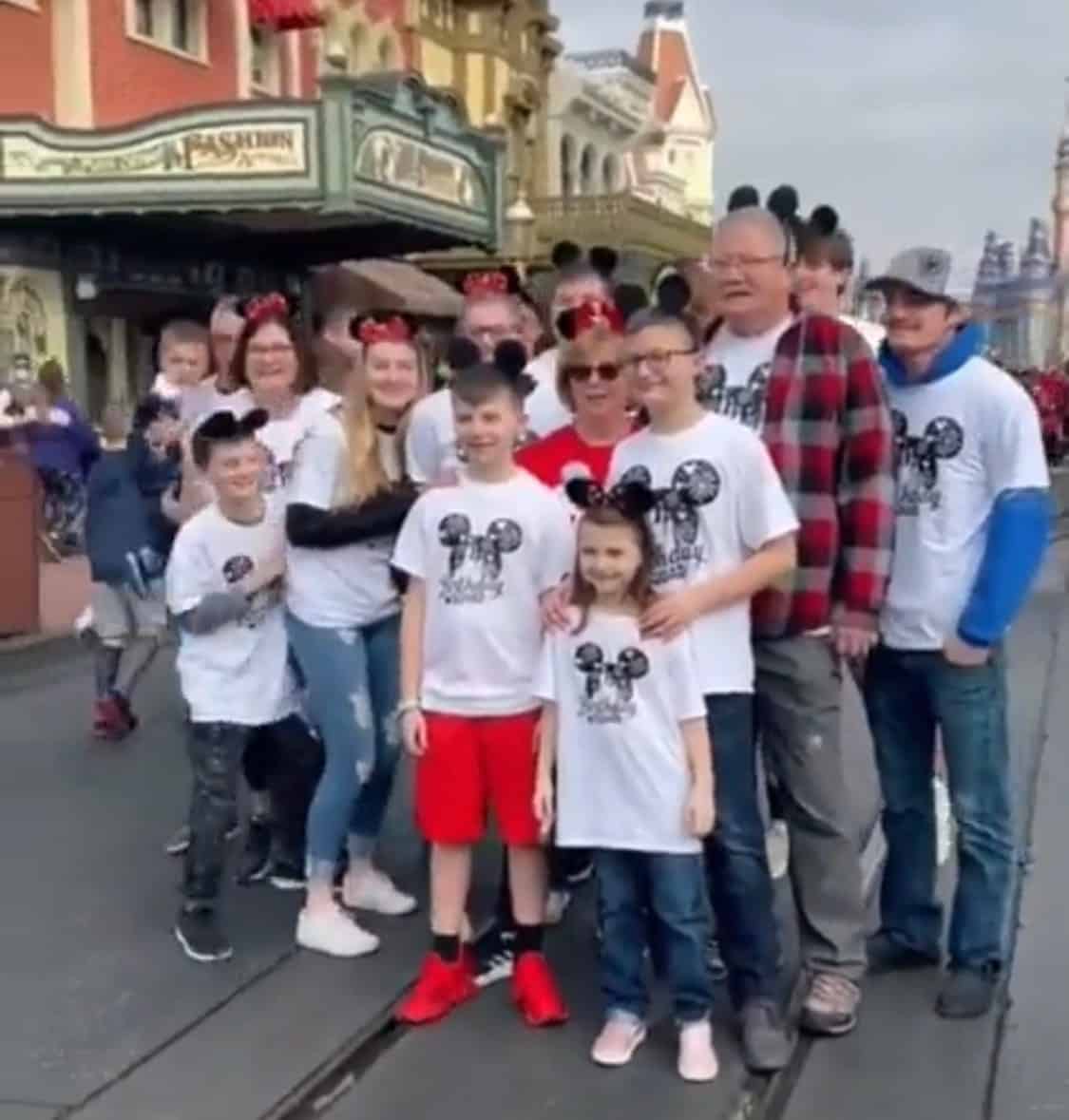 A family decked out in matching Mickey Mouse shirts and ears gathers on Main Street, U.S.A. at Disney World, recording a joyful surprise video as they celebrate their loved one’s 70th birthday with a magical reveal.