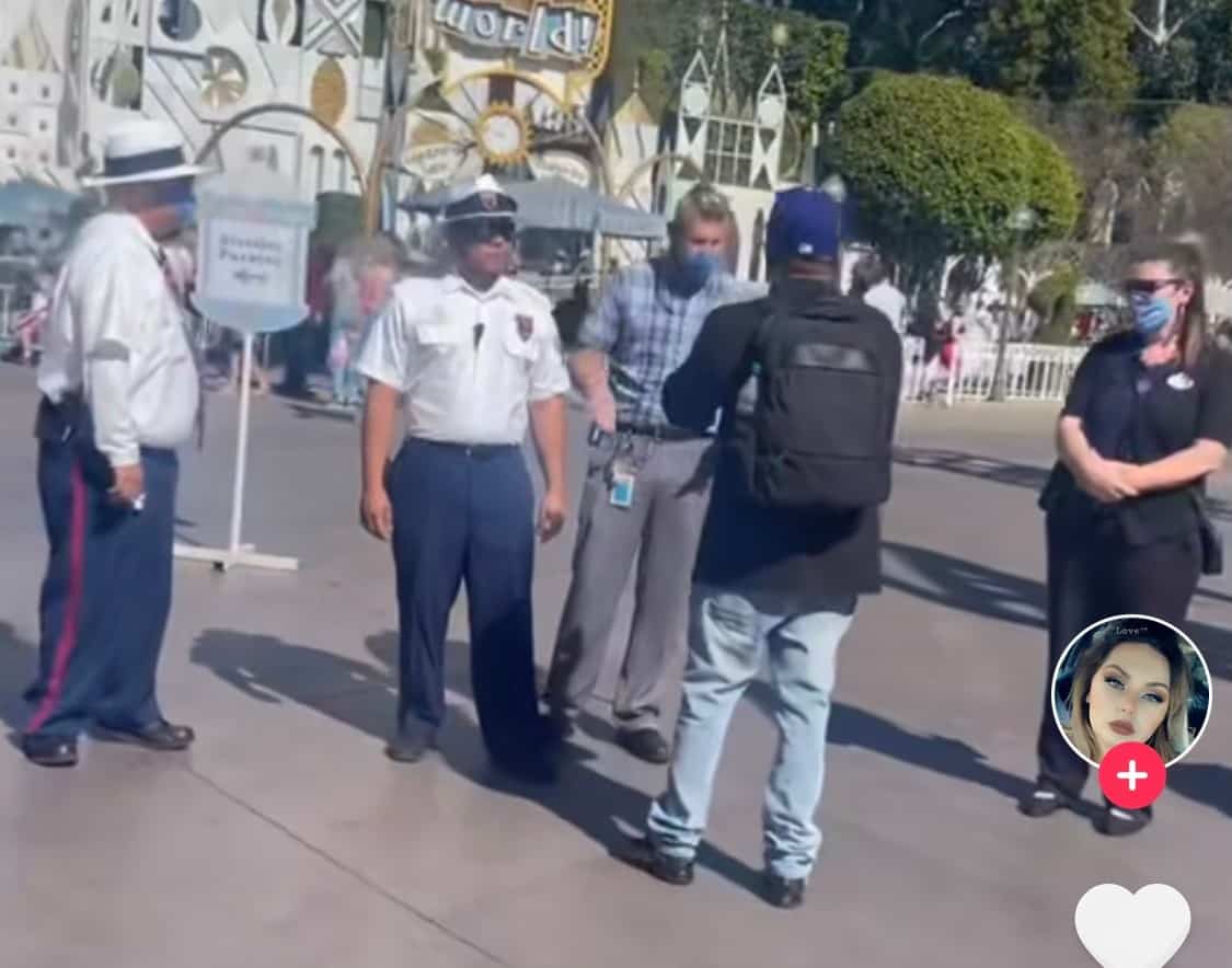 Disneyland security staff talk with a guest in a black shirt and backpack at the park entrance—looks like another tense moment unfolding right outside the Magic Kingdom gates.