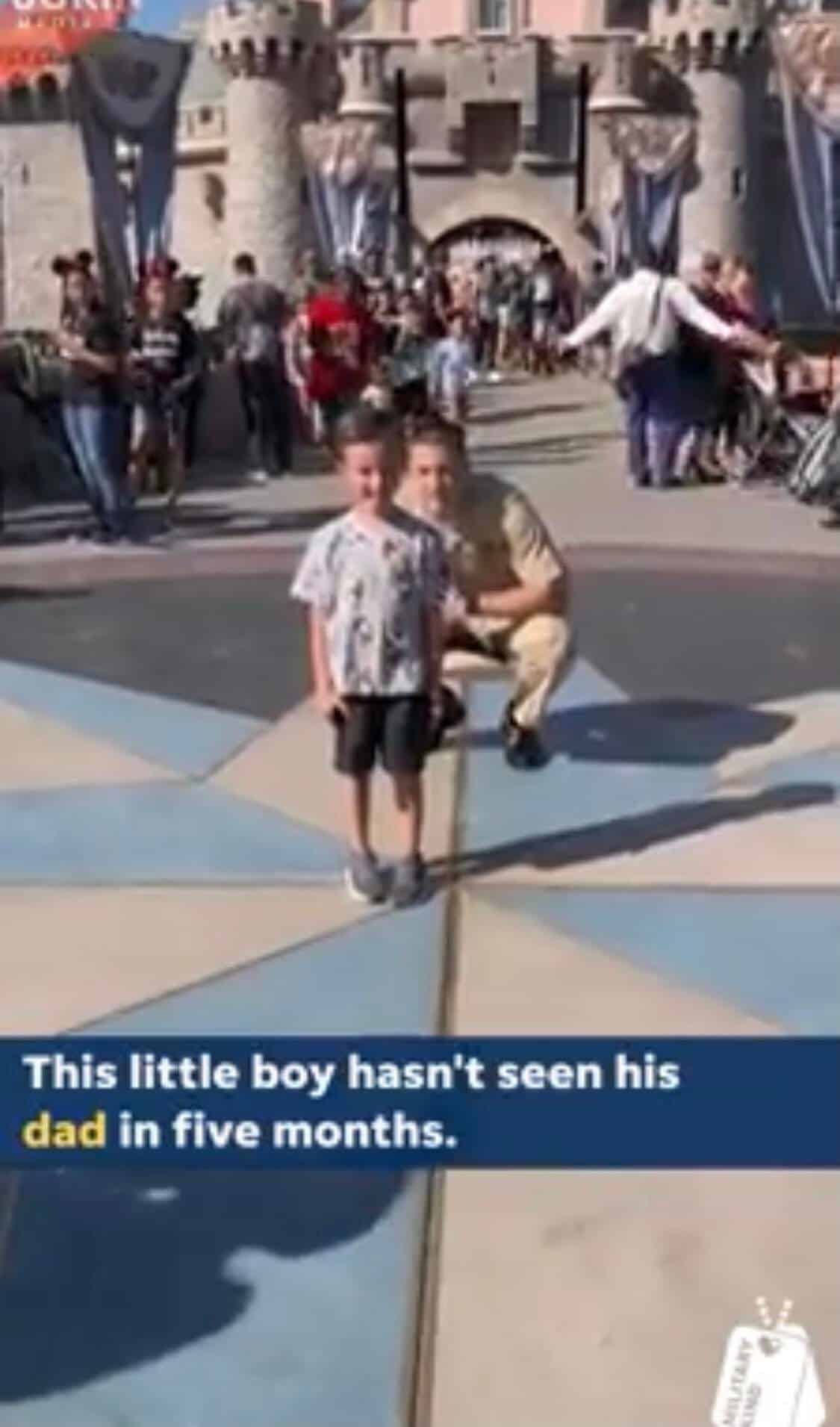 A smiling boy beams with excitement as an adult kneels behind him at the bustling Disneyland entrance, capturing a magical moment as he receives an unforgettable surprise in this touching video.