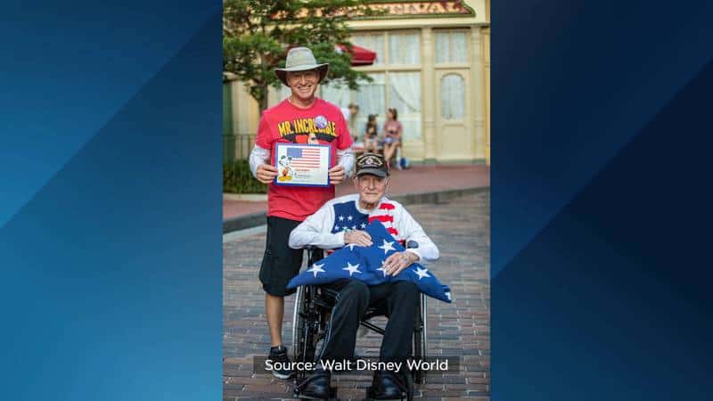 At Walt Disney World, a WWII veteran marks his 100th birthday in a wheelchair while a smiling boy stands behind him, proudly holding a folded American flag amid the park’s festive atmosphere.