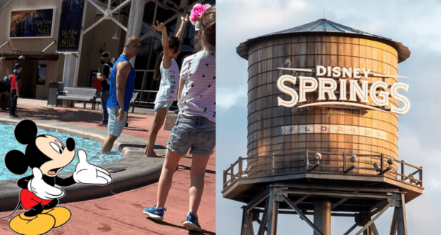 Mickey Mouse happily waves to children by a sparkling fountain, with the iconic Disney Springs water tower in the background. Nearby, a guest stands just beyond the marked area, seeming to ignore the park guidelines.