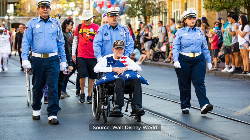 A WWII veteran rides in a wheelchair, proudly holding an American flag, as Disney cast members and uniformed officers escort him down Main Street, U.S.A. in a festive parade at Walt Disney World, celebrating his 100th birthday while cheerful crowds look on.