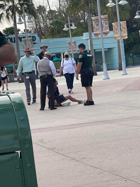 A guest is seated on the ground at Disney’s Hollywood Studios, surrounded by security and staff, with palm trees visible in the background—a scene reminiscent of recent viral moments involving a woman in handcuffs at the park.