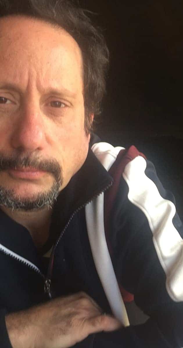 A theme park enthusiast with short dark hair and a goatee, dressed in a zip-up jacket, gazes thoughtfully into the camera amid widespread news in the entertainment world of David Brenner’s passing at 59.