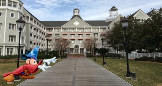Mickey Mouse dressed as a wizard welcomes guests along a walkway leading to a grand gray hotel with white trim at Walt Disney World, highlighting the new Interactive TVs now available as part of your Disney resort stay.