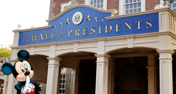Mickey Mouse stands outside the Hall of Presidents at Disney World, playfully gesturing for silence, as Cast Members hope guests will enjoy the show without turning it into a political discussion.
