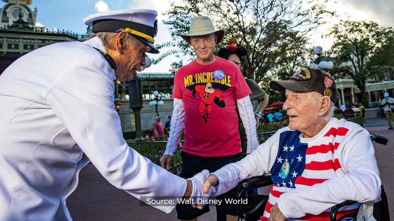A uniformed cast member warmly shakes hands with a cheerful WWII veteran seated in a wheelchair, with another guest standing nearby. The festive moment unfolds as the veteran celebrates his 100th birthday at Walt Disney World, surrounded by magical atmosphere and joyful smiles.