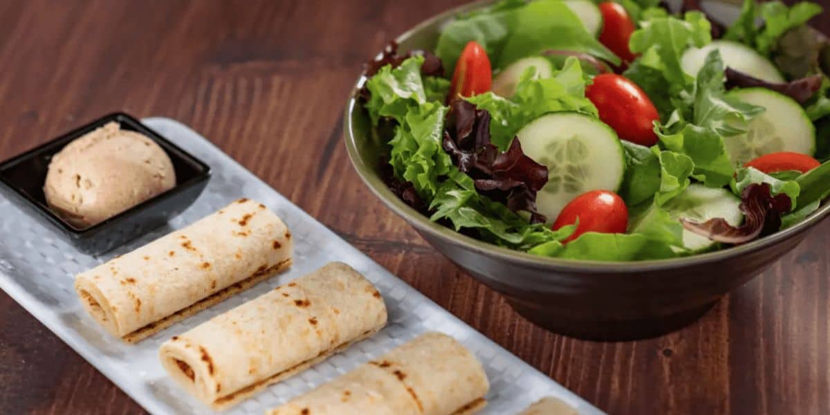 A fresh salad with mixed greens, cucumber slices, and cherry tomatoes sits in a bowl, perfect for healthy eating. Next to it, three rolled flatbreads lie on a rectangular plate with a small dish of dipping sauce. It's like dining at Walt Disney World right in your own home!