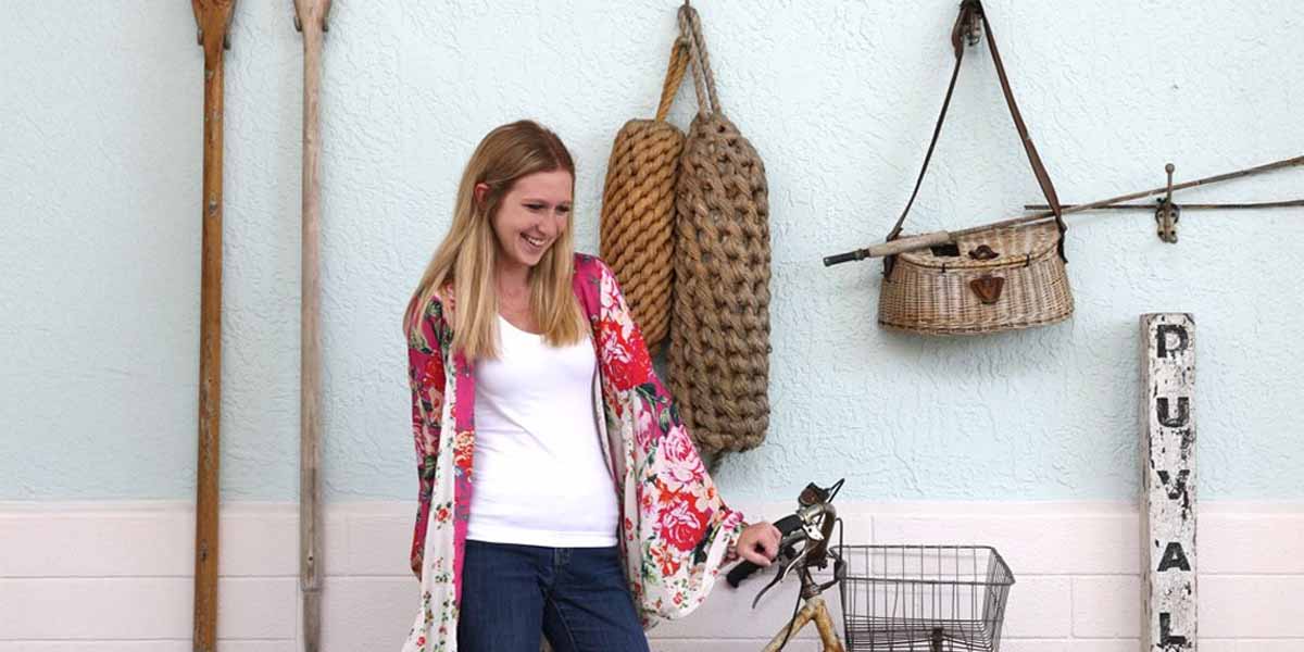 A woman with long blonde hair, wearing a white shirt and floral kimono, smiles while standing next to a vintage bicycle against a light blue wall adorned with nautical items inspired by Disney Resort Decor.