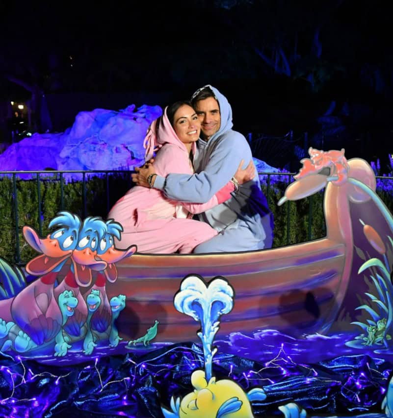 Guests in casual hoodies share a warm embrace aboard a whimsical cartoon boat attraction, surrounded by lively animated blue water and playful sea creatures—bringing to life the fun, lighthearted atmosphere reminiscent of John Stamos making a surprise Disney appearance.