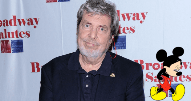 A man in a navy blazer stands in front of a Broadway-inspired backdrop featuring Mickey Mouse, as fans and the entertainment community honor legendary designer Tony Walton following news of his passing at 87.