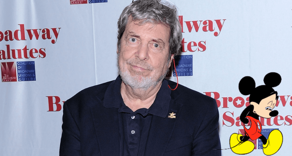 A man in a navy blazer stands in front of a Broadway-inspired backdrop featuring Mickey Mouse, as fans and the entertainment community honor legendary designer Tony Walton following news of his passing at 87.