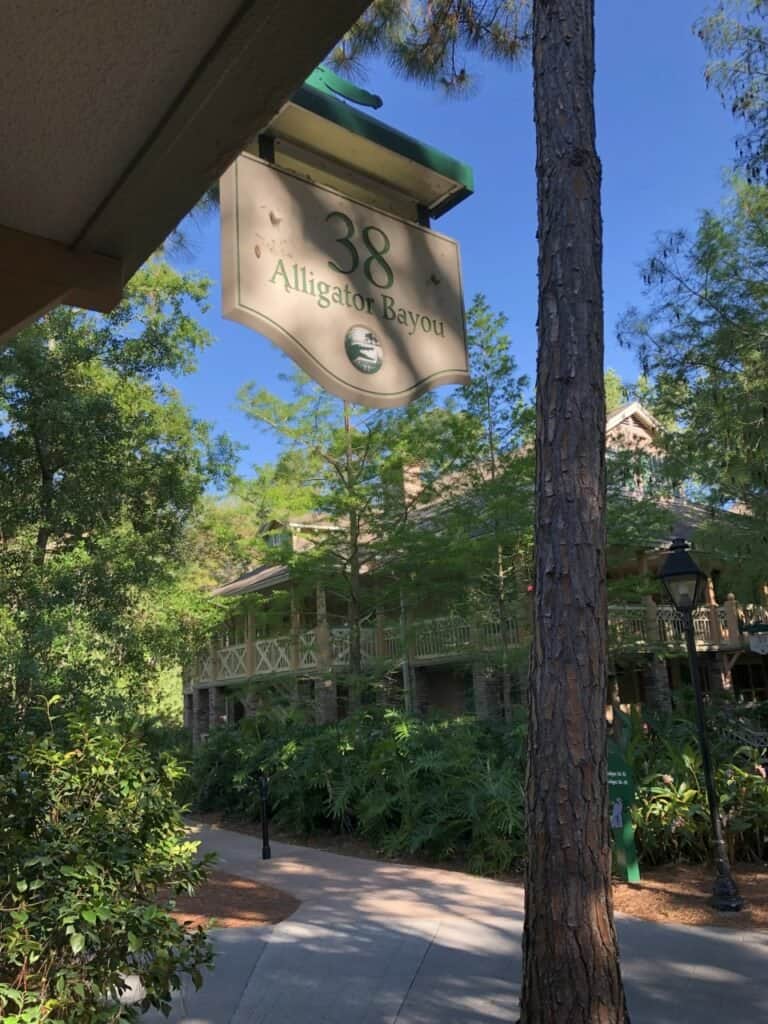 The "38 Alligator Bayou" sign welcomes guests amid lush trees and picturesque buildings at Disney’s Port Orleans Riverside Resort, set against a bright, sunny sky.