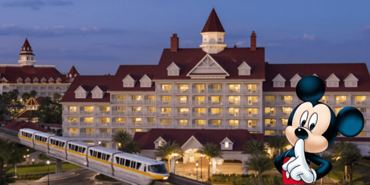 At dusk, a large deluxe hotel with a red roof and white exterior glows with warm lighting. A monorail glides past, embodying the charm of a Disney Resort. In the foreground, an animated character stands thoughtfully with a finger to their mouth.