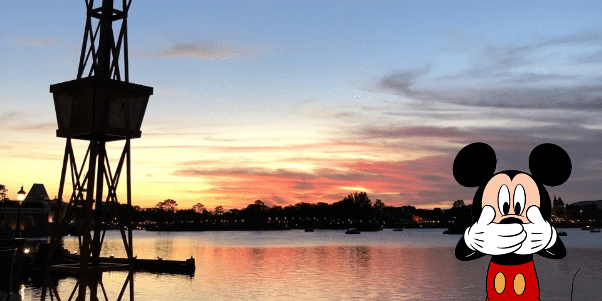 Mickey Mouse playfully covers his mouth while standing beside the serene Bay Lake at sunset, with iconic silhouettes of Disney World’s attractions and lush trees in the background, creating a magical Disney moment.